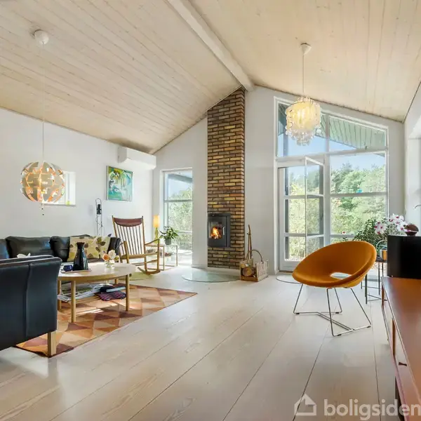 Stue med sofa, pejs og gyngestol i handling. Store vinduer viser grønne omgivelser. Trægulve og loft skaber en lys og rummelig atmosfære, kompletteret med moderne møbler og udsmykning.