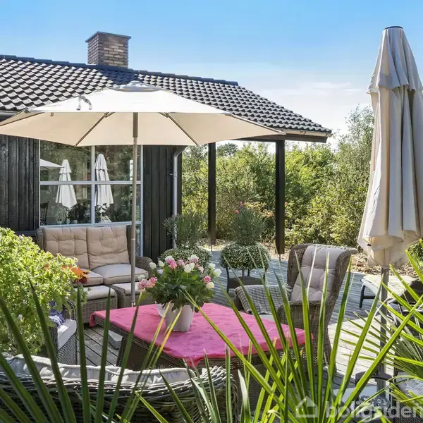 En terrasse med havemøbler under en hvid parasol. Møblerne omfatter en sofa og stole, omgivet af frodige grønne planter. I baggrunden ses et mørkt træhus.