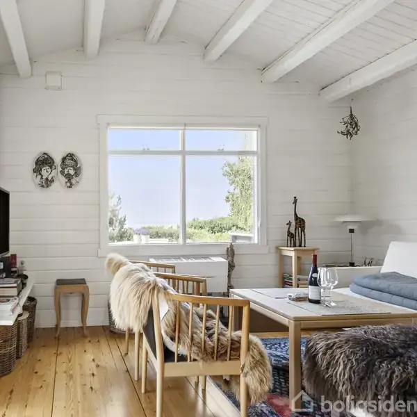 Stue med træmøbler dækket af pels, en sofa og et bord med en vinflaske. Et stort vindue viser udsigt til natur. Hvidmalede vægge og træloft giver en lys atmosfære.