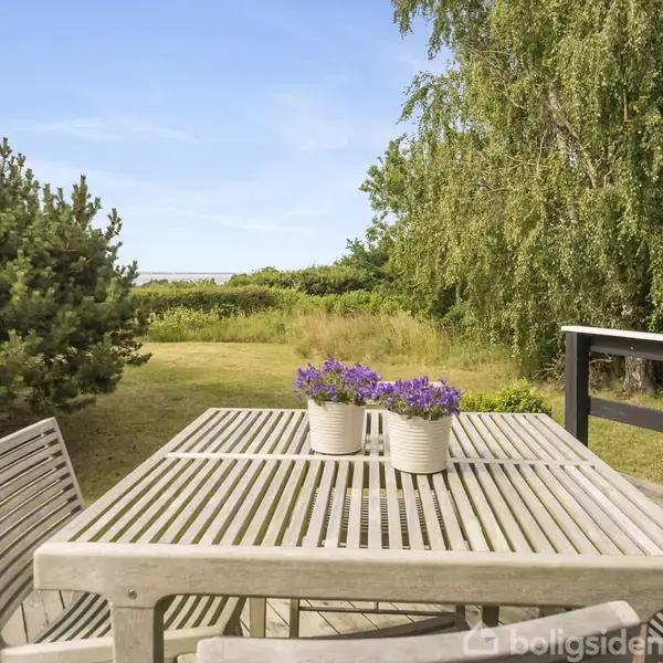 Bord med stole står på en terrasse med udsigt til en græsplæne omgivet af træer og buske. To blomsterpotter med lilla blomster er placeret på bordet.