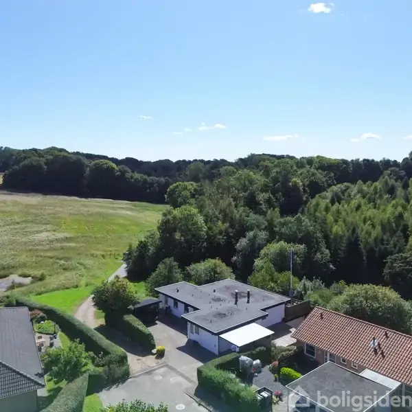 Hus ligger stille mellem andre huse omgivet af tæt bevoksede områder og en stor mark, under en klar blå himmel.