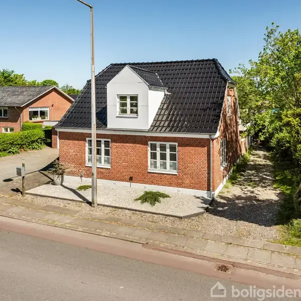 Et rødt murstenshus med sort tag står ved en vej, omgivet af grønne træer og buske i et beboelseskvarter.