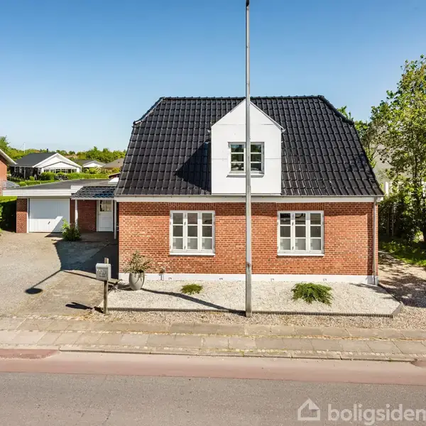 En rød murstensvilla står stille på en solrig dag omgivet af en forhave med stiklinge og en tilstødende garage. Villakvarteret er roligt med grønt løv omkring.