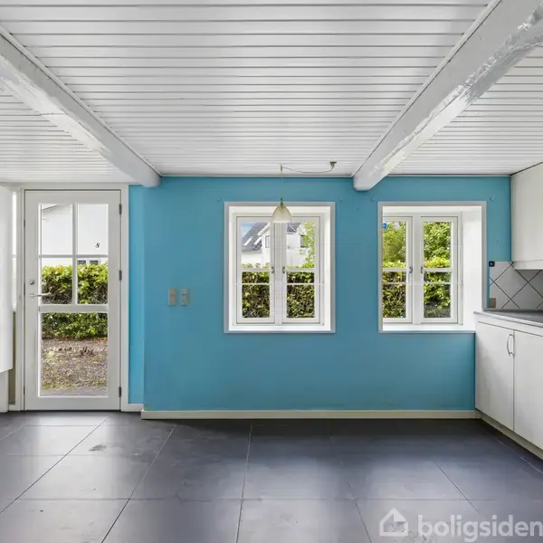 Et køkken med hvide skabe og blå vægge. Vinduet og dør åbner til en have. Der er en hvid loftslampe og sorte gulvfliser.