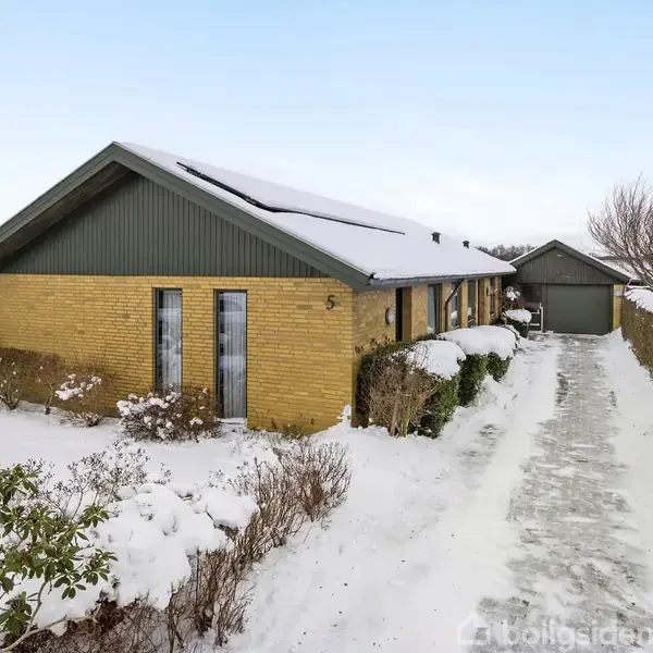 Gul murstenshus dækket af sne står stille i et vinterlandskab med en lang indkørsel, flankeret af snedækkede buske, der fører til en garage.