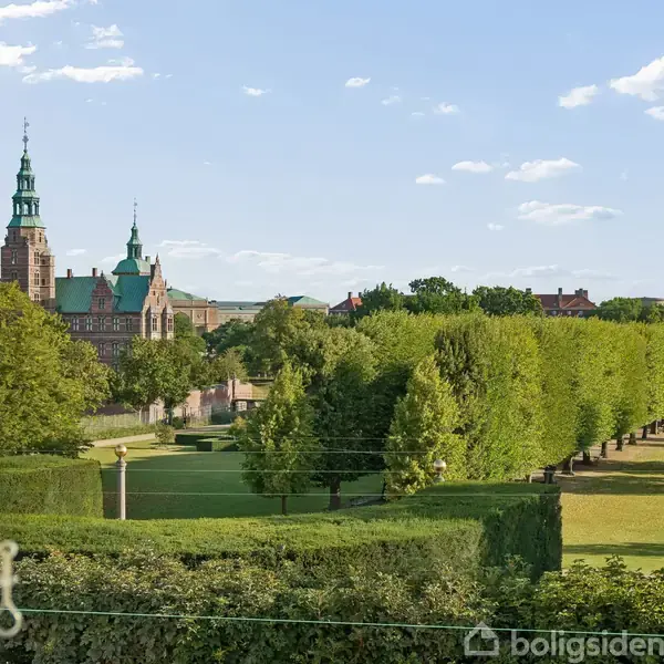 Slot med tårne ses fra et åbent vindue med en udsigt over en velplejet have med trimmede planter og en allé af træer. Himlen er blå med lette skyer.