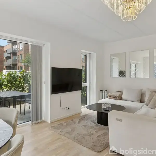 Stue med lys sofa arrangeret rundt om et sofabord, væghængt fladskærms-tv, der vender mod et åbent altanområde med havemøbler, omgivet af grønne buske og boligbygninger.