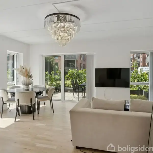 En lys stue med en beige sofa står foran et spejl og vægmonteret tv. Rundt spisebord ved vinduer, der åbner til en balkon med udsigt over grøn beplantning.