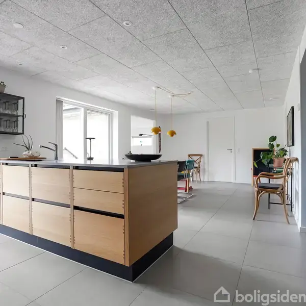 Et moderne køkken domineret af en lys træ-køkkenø med sort bordplade. I baggrunden ses en spiseplads med stole, grønne planter og store vinduer, der giver naturligt lys.