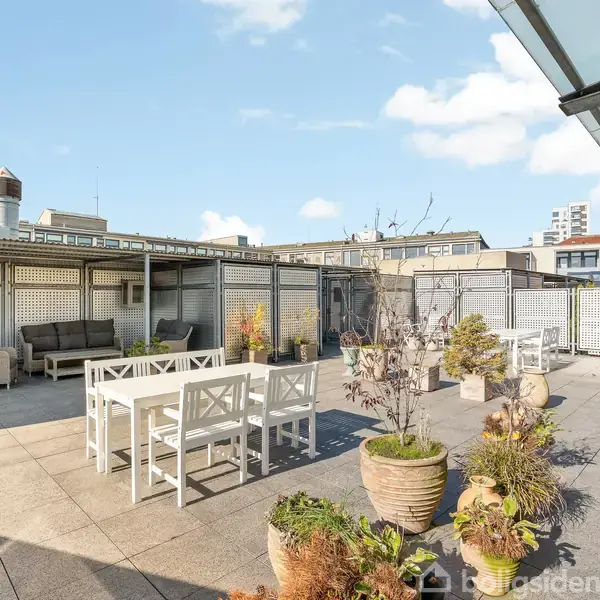 Terrasse med hvide havemøbler ved spisebord; planter i krukker på fliserne. Overdækket loungeområde med sofa til venstre. Bygninger og blå himmel med få skyer i baggrunden.