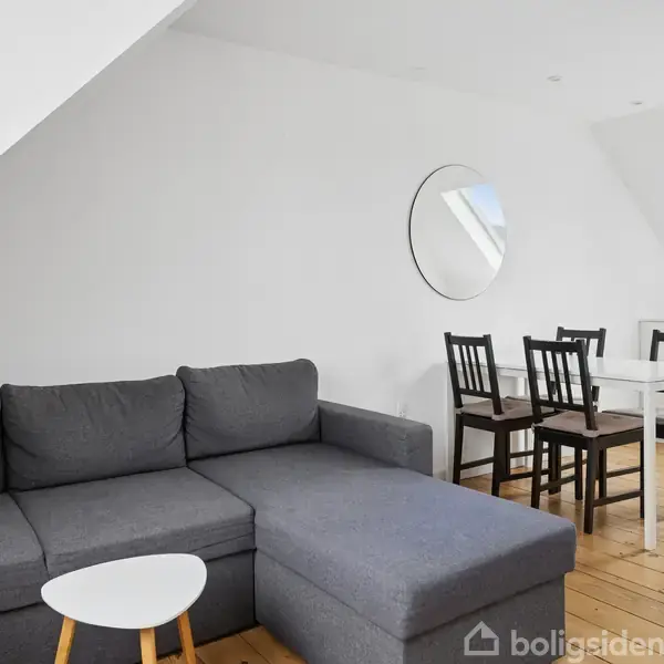 Grå sofa står ved siden af et rundt, lille bord i en stue. I baggrunden ses et spisebord med stole. Loftet har skrånende vægge og ovenlysvinduer.