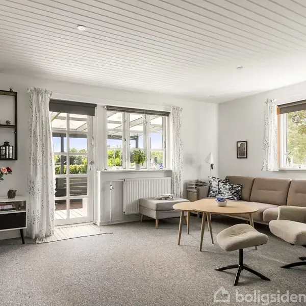 En lys stue med en beige sofa og lænestol, placeret omkring et lille træbord, har udsigt til en terrasse gennem glasdøre. En reol med dekorationer står til venstre.