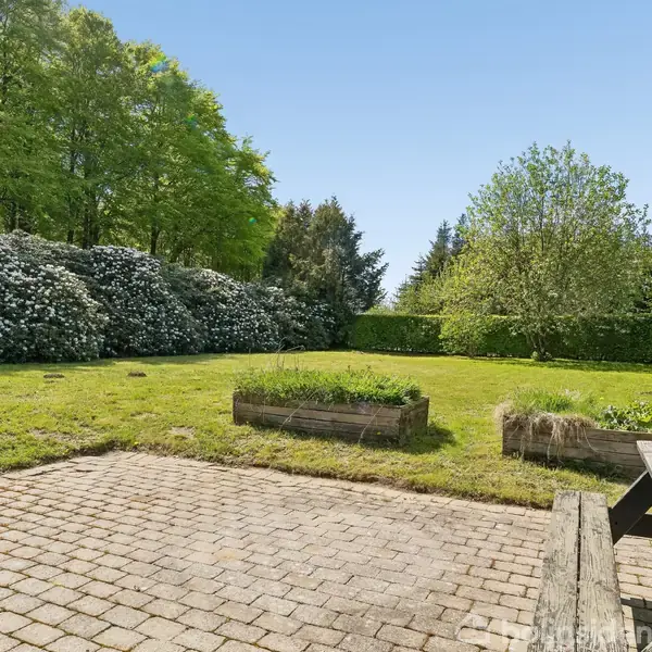 Træbord står på en brolagt terrasse, omkranset af grønne buske og træer i en have. En græsplæne med plantekasser ses i baggrunden under en klar blå himmel.