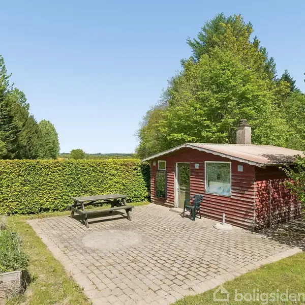 Et rødt træhus står i en frodig have omkranset af hække. Foran huset er en terrasse med et picnicbord og stole. Omkring huset er der tætte grønne træer.