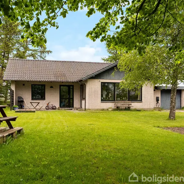 Et enfamiliehus i gule mursten, med gråbrunt tegltag, i en grøn have med træer. En terrasse med en træbænk og et bord står til venstre i billedet.