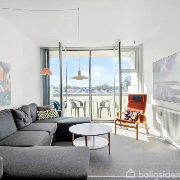 En grå hjørnesofa står langs væggen med kunst på. I en lys stue med åbne altandøre er en orange lænestol. På altanen ses stole og bord.