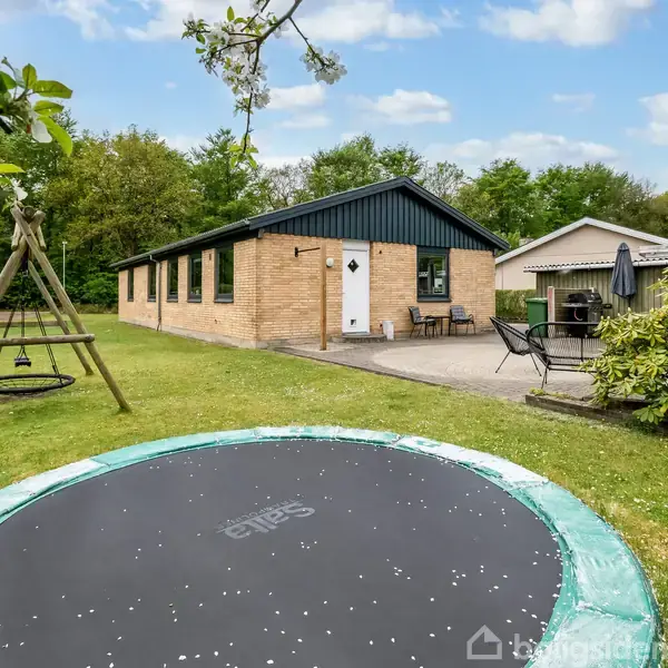 Trampolin er placeret i en græsplæne foran en gulstensbygning med sort tag. Til højre står et gyngestativ, og der er en terrasse med havebord og stole.