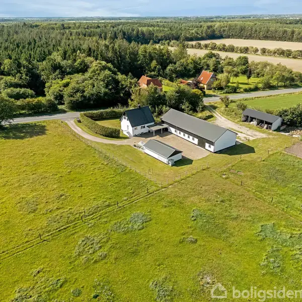 Hus omgivet af grønne marker og skov. Det hvide hus med et sort tag ligger ved en vej, sammen med en stor bygning og en mindre struktur.