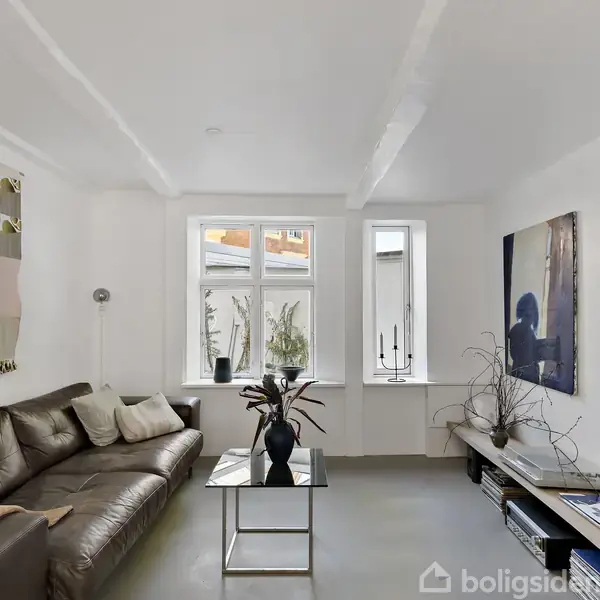 En brun lædersofa står langs en væg med moderne kunst. Et glasbord med en vase står foran. Vinduet bagved giver lys, og vægdekorationer tilføjer farve til rummet.