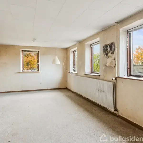 Et tomt rum med fire vinduer tillader naturligt lys at strømme ind. Væggene er slidte med tapet, der skræller af, og en radiator står under vinduerne.