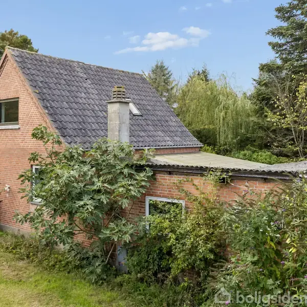 Et rødt murstenshus omgivet af frodig vegetation, med en skråt tag og en skorsten. Huset har vinduer på begge etager. Omgivet af træer og buske i en have.