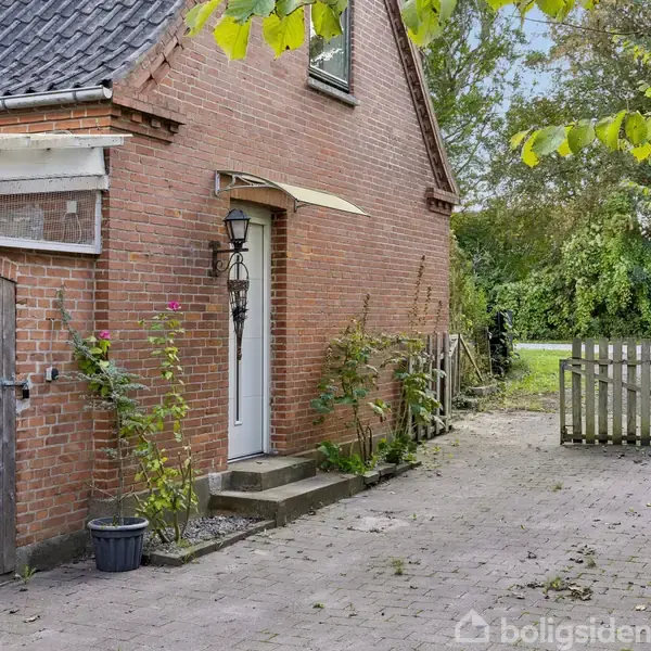 Murstenshus med sort tegltag, omkranset af en flisebelagt indkørsel. En lanterne hænger ved hoveddøren. Planter vokser langs facaden. I baggrunden ses en grøn have og en trælåge.
