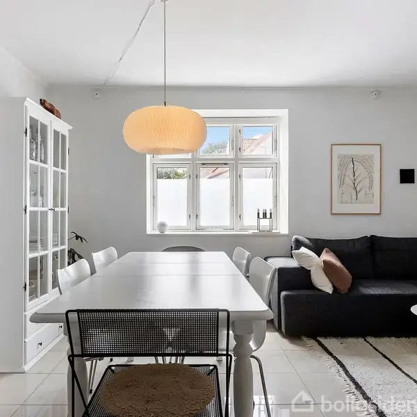 Spisebord står i et lyst rum med hvide vægge og lyse fliser. Rundt om bordet er der stole, en sort sofa og en reol. En stor lampe hænger over bordet.