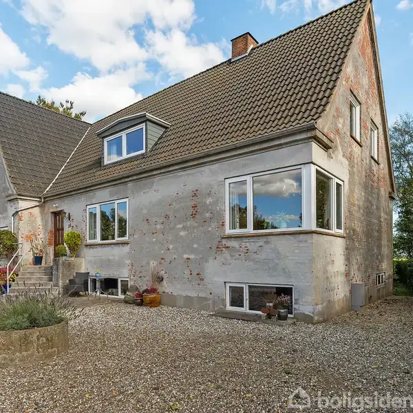 Et to-etagers hus med grå facade står stille på en grusbelagt grund. Omgivet af en lav hæk og buske under en blå himmel med spredte skyer.