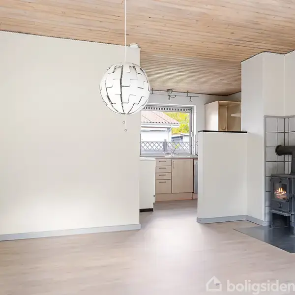 En stue med en moderne, hvid hængelampe, et brændeovnshjørne og træloft, åbner sig mod et lyst køkken med vindue. Enkel, minimalistisk indretning.