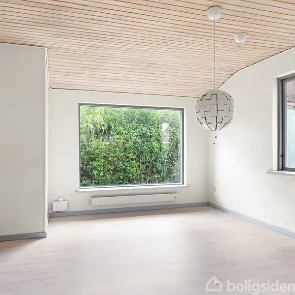 Et lyst rum med trægulv og træloft har en moderne lampeskærm hængende. Store vinduer giver udsigt til tæt grønt buskads udenfor.