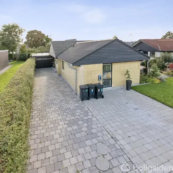 Et parcelhus med et fladt tag og gul murstensfacade står ved siden af en flisebelagt indkørsel. Tre affaldscontainere er placeret tæt på huset. Græsplæne og hække omgiver området.