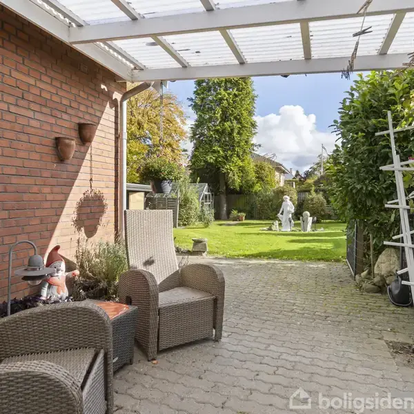 To fletstole og et bord på en flisebelagt terrasse under overdækning. I baggrunden ses en grøn græsplæne med en hvid statue og farverige træer.