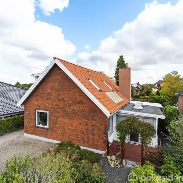 Rødstenshus med rødt tag står stille omgivet af en grøn have og nabohuse. Der er en grusbelagt indkørsel ved siden af huset. Skyer dækker delvist den blå himmel.