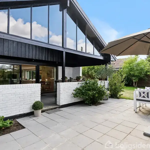 Et moderne hus med store glasvinduer og hvid murstensfacade. Foran huset er en terrasse med havemøbler under en parasol, omgivet af grønne planter og en græsplæne i baggrunden.