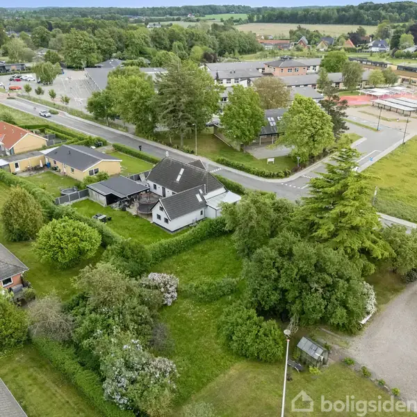 Huse med haver ligger langs en vej omgivet af træer og grønt område. En skole eller offentlig bygning kan ses i baggrunden.
