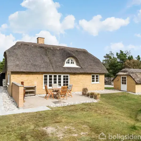 Stråtækt hus står roligt med træmøbler på terrassen, omgivet af græs og klitter. Et mindre stråtækt anneks ses tæt ved. Skoven danner baggrunden under en blå himmel med skyer.