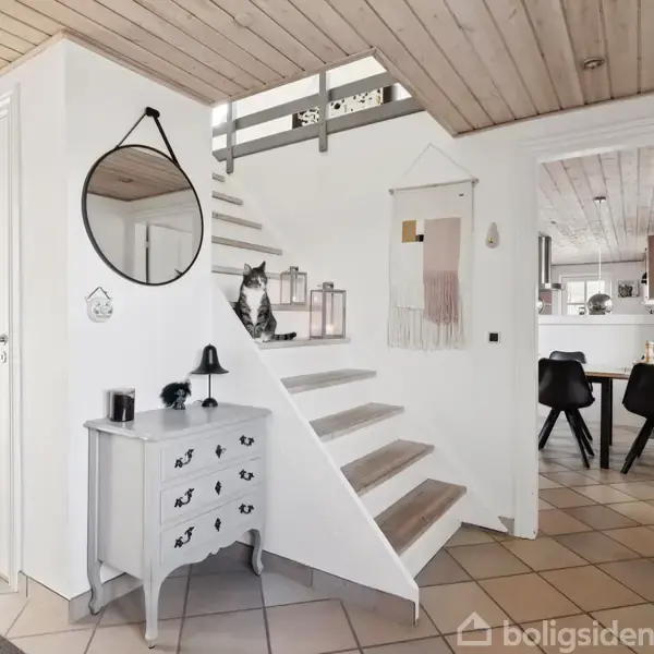 Kattefigur sidder på en hvid trappe, flankeret af lanterner. Et gråt bord står foran trappen, dekoreret med lampe og spejl på væggen. Til højre ses spiseområde.