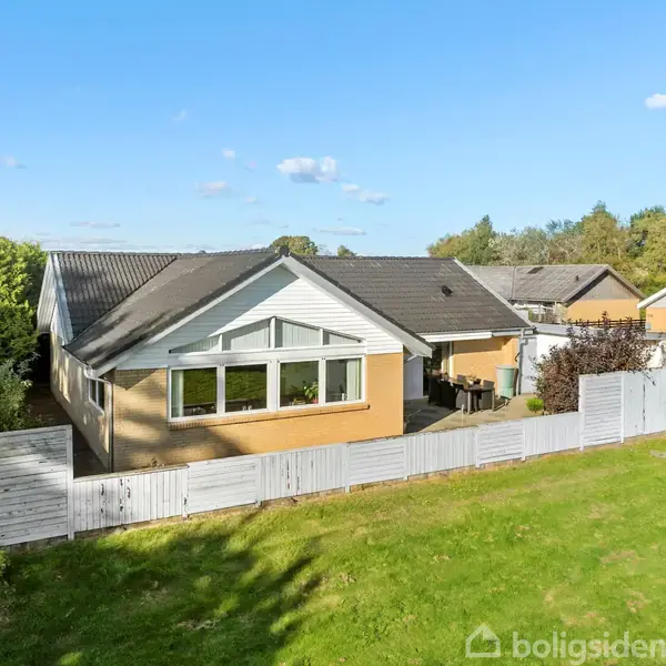 Hus med gavl, omkranset af et hvidt stakit, står i en grøn have. Bag det ses andre boliger og træer under en blå himmel med få skyer.