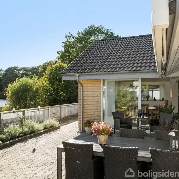 Et udendørs terrasseområde har et bord og stole med planter omkring. Det er solrigt, omgivet af buske og træer langs en havemur.