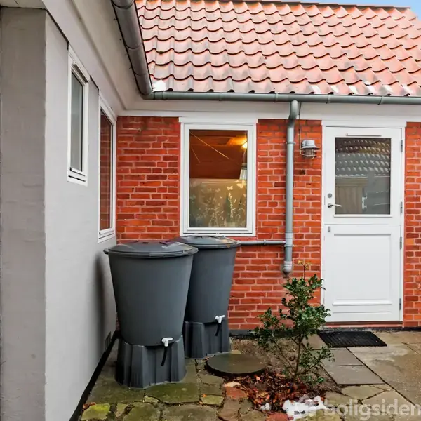 Et rødt murstenshus med et stisystem af fliser og to regnvandstønder. En hvid dør og et vindue er synlige. Et lavt plankeværk grænser haven.