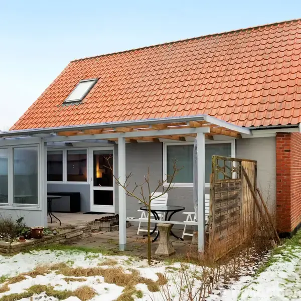 Huset har et rødt tegltag og store vinduer. Der er en overdækket terrasse med stole og et bord i en vinterlig have med snepletter omkring.