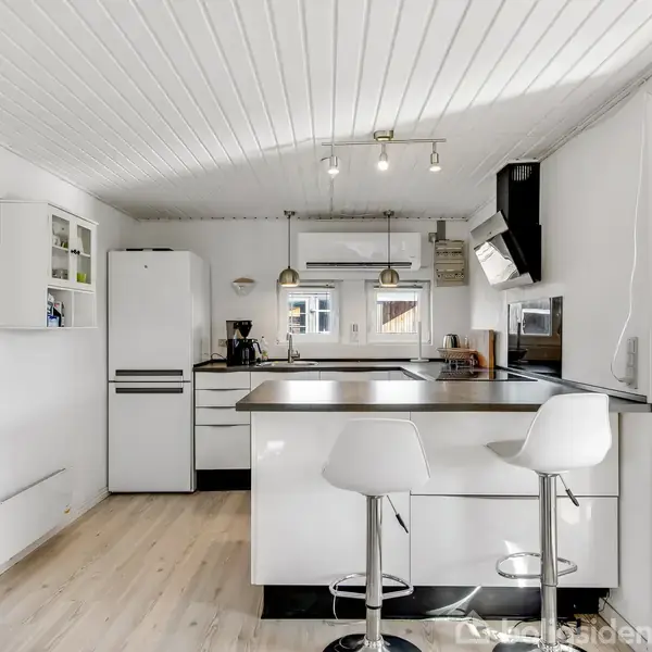 Hvidt, moderne køkken med køleskab og kogeplade, to barstole ved køkkenø. Lyse trægulve og hvide vægge med et lille maleri. Stor vinduesdør leder ud mod terrasse.