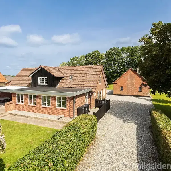 Huset er en rød murstensbygning med et skråt tag og en udendørs terrasse. Det står i en have med grusvej, omgivet af buske og et fritstående rødt udhus i baggrunden.