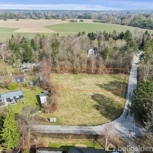 Græsmark afgrænset med orange linjer ligger i et kvarter med spredte huse og tæt skov. Veje snor sig gennem området, og marker er synlige i baggrunden.
