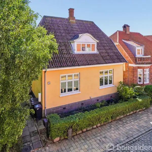 Et gult hus med mansardtag og en stor kvist står ved siden af et rødt murstenshus. Foran er en flisesti og en smal hæk. En skyggefuld vej er synlig.