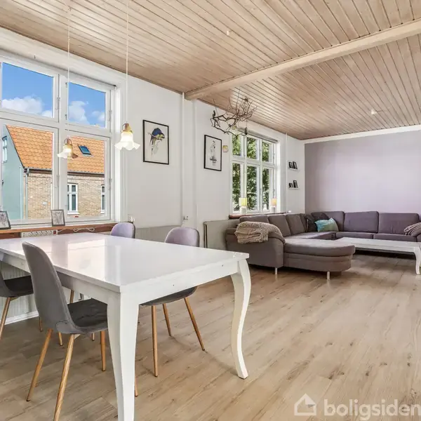 En spisestue og stue med spisebord og stole ved vinduet, der lukker naturligt lys ind. I baggrunden er en grå hjørnesofa med puder. Trægulvet skaber en varm atmosfære.