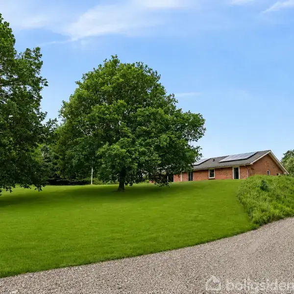 Hus beliggende på en gruset vej omgivet af grønne træer og græsplæne under en klar blå himmel.