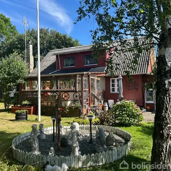 Rødt hus med veranda, omgivet af have med græs og træer. I forgrunden er der en rund stenbed dekoreret med figurer og en flagstang. Solrig dag med blå himmel.