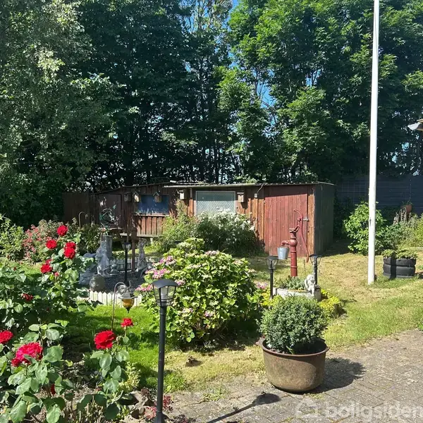 En velplejet have med blomsterbede og græs, dekoreret med røde blomster og buske. En flagstang står ved siden af et træhus i baggrunden, omgivet af grønne træer.