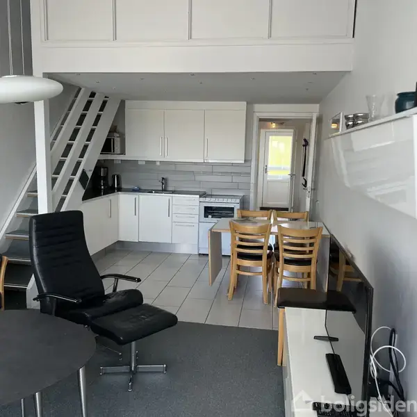 Stue med sort lænestol, sofabord og tv. Køkkenet i baggrunden med spisebord og stole. Trappe til højre fører til en hems eller første sal.